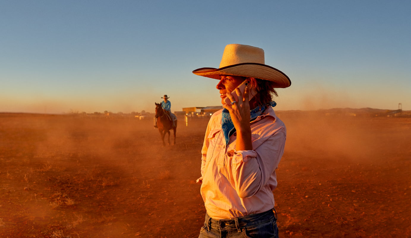 A farmer wearing an Akubra holds a mobile phone to their ear and is smiling. They are surrounded by desert with outlines of a farm in the distance.