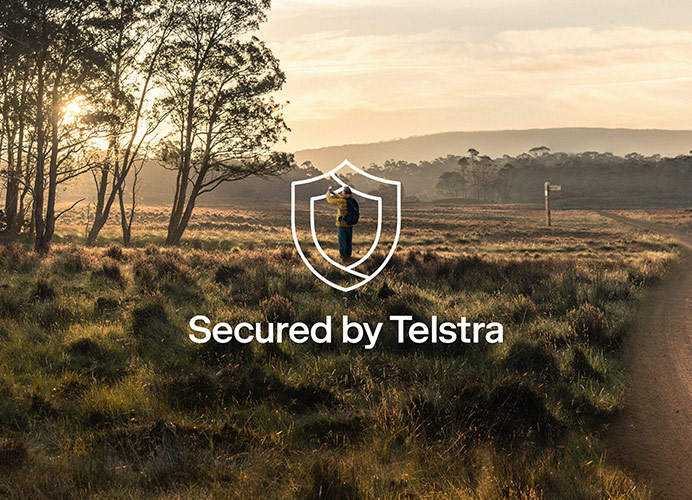 A man standing in a field by a country road, taking a picture of the sky. A bold text overlay shows a shield and is accompanied by the words: Secured by Telstra.