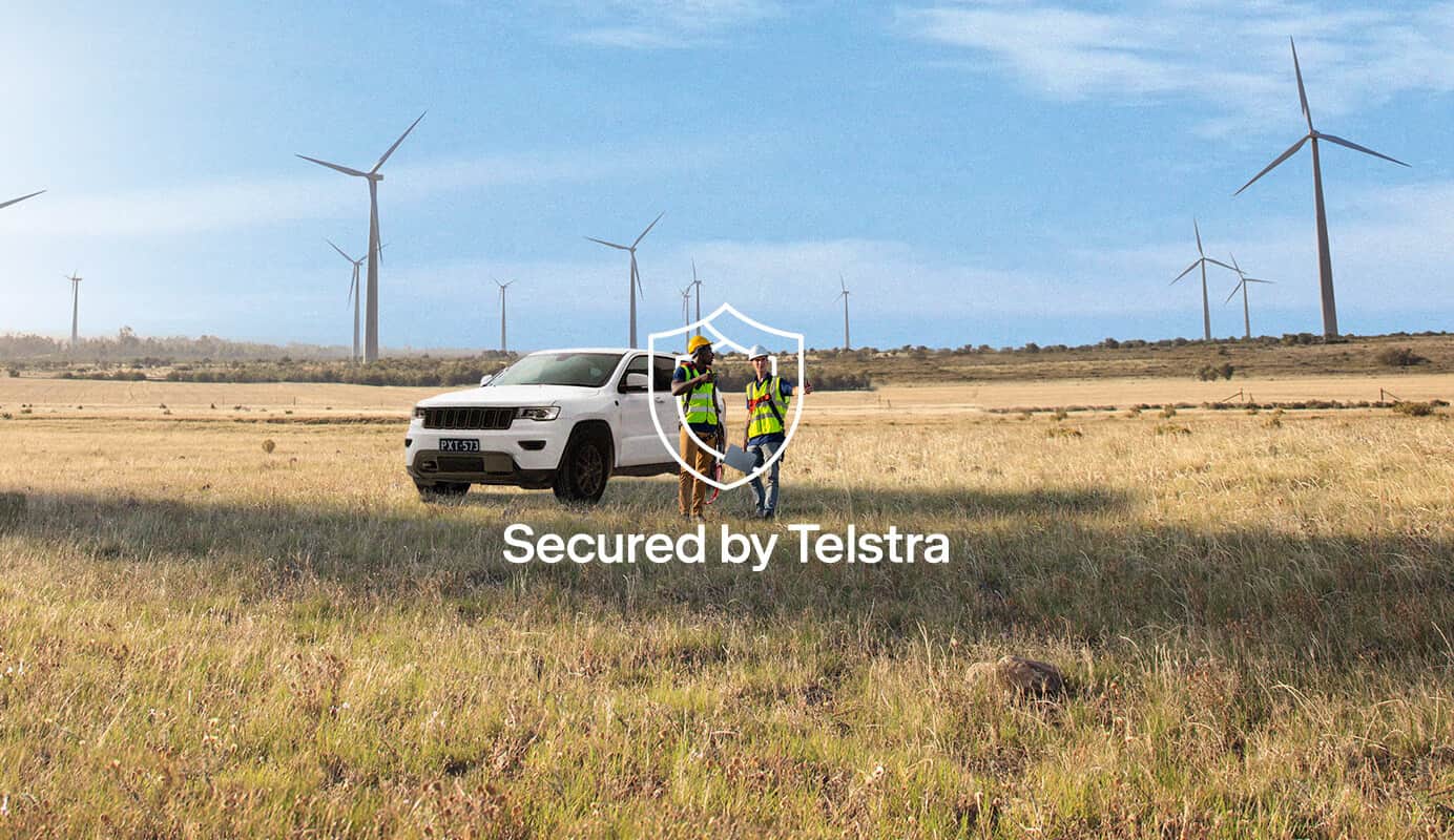 Two people standing in front of a white four-wheel drive in a regional area with windmills in the background. Text on image: Secured by Telstra