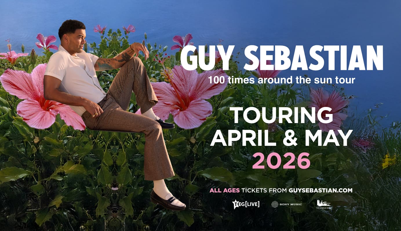 Guy Sebastian sits among vibrant pink flowers under a clear blue sky, featured on the official poster for his 100 Times Around The Sun tour. Text highlights the tour dates in April and May 2026. All ages tickets from guysebastian.com