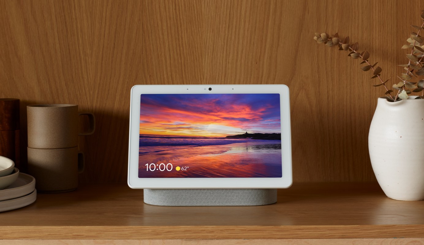 A Google Nest Hub Max rests on a side table displaying time of day, the weather and a beautiful, sharp sunset image as wallpaper.