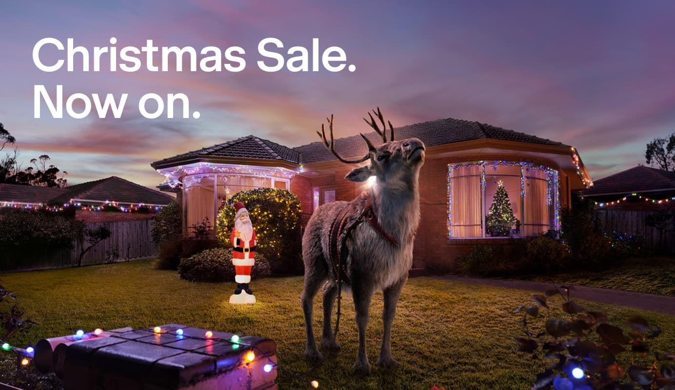 A lost reindeer stands in the front yard of a suburban house looking wistfully into the sky. The house is lit up with Christmas decorations. Text on image: Christmas Sale. Now on.