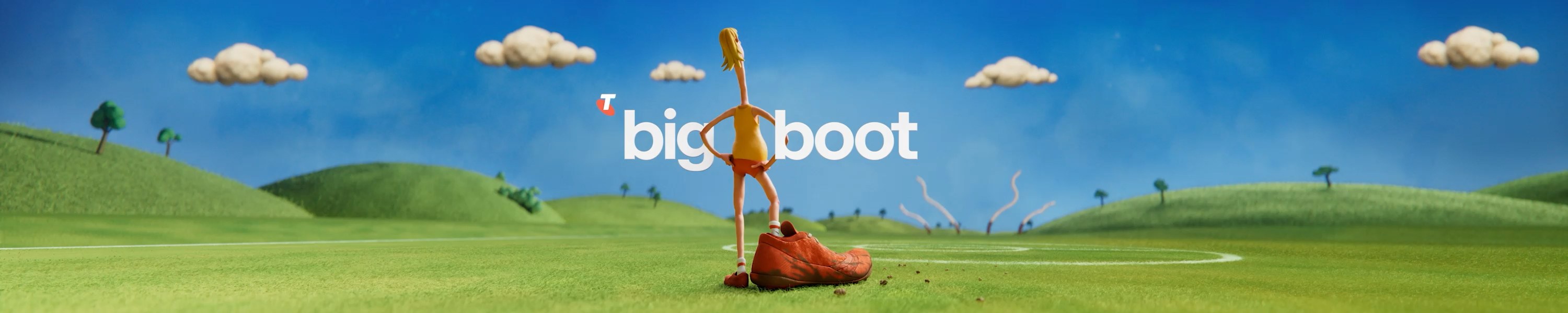 A person dressed in a yellow non-descript Australian Football League guernsey with orange footy shorts. The person is wearing orange football boots, with the right boot being significantly larger than the left for comedic effect and to also reinforce the promotion name ‘Big Boot’. They are sitting on giant letters that form the word ‘Big Boot’ in a grass field under a clear sky.