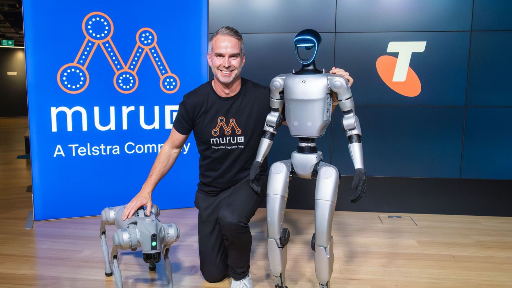 Ben Green kneels between a robotic dog and humanoid robot, with muru-D and Telstra logos on screens behind.