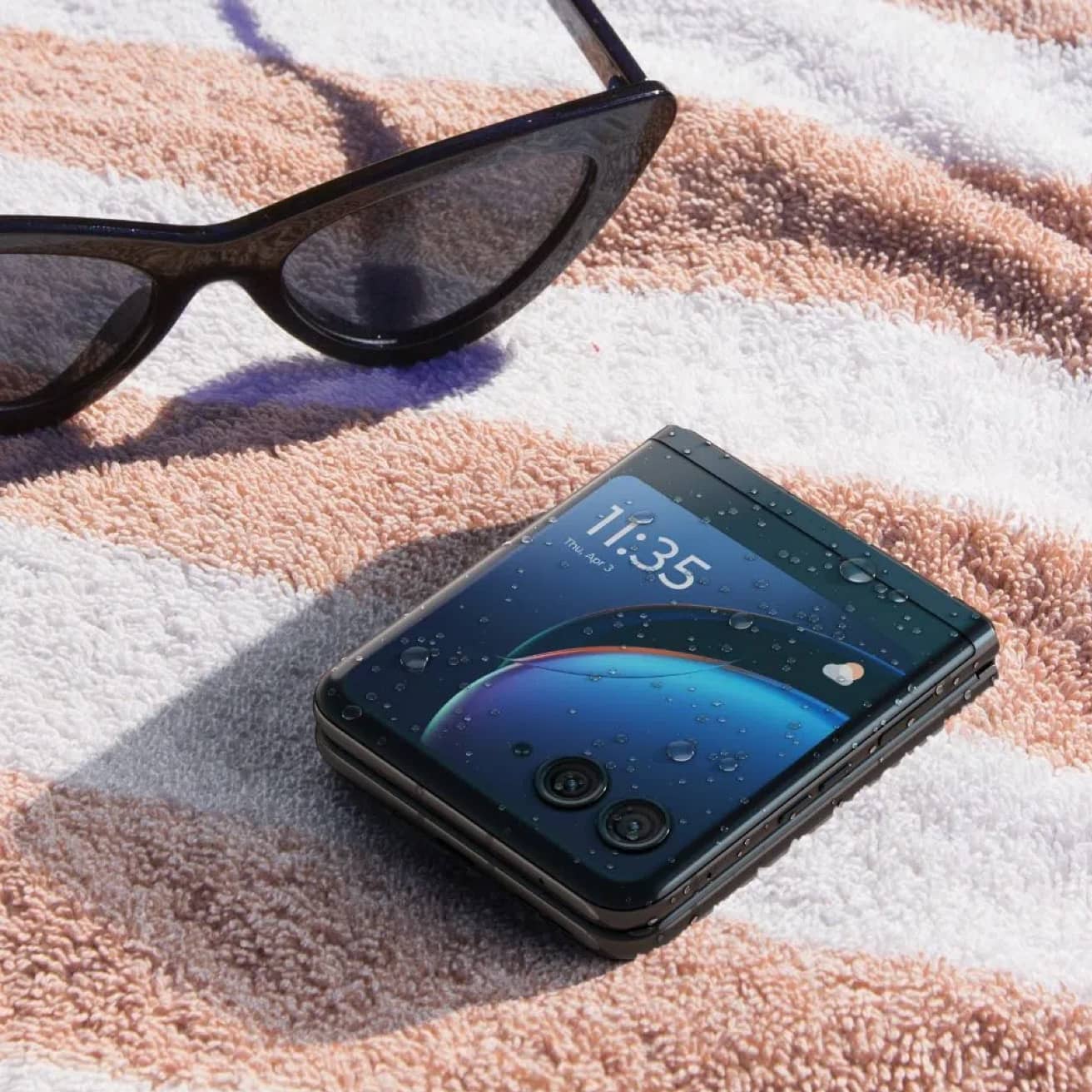 A closed motorola razr 40 ultra rests on a beach towel, covered with water droplets. The external display depicts the default home screen, indicating the phone is still working.