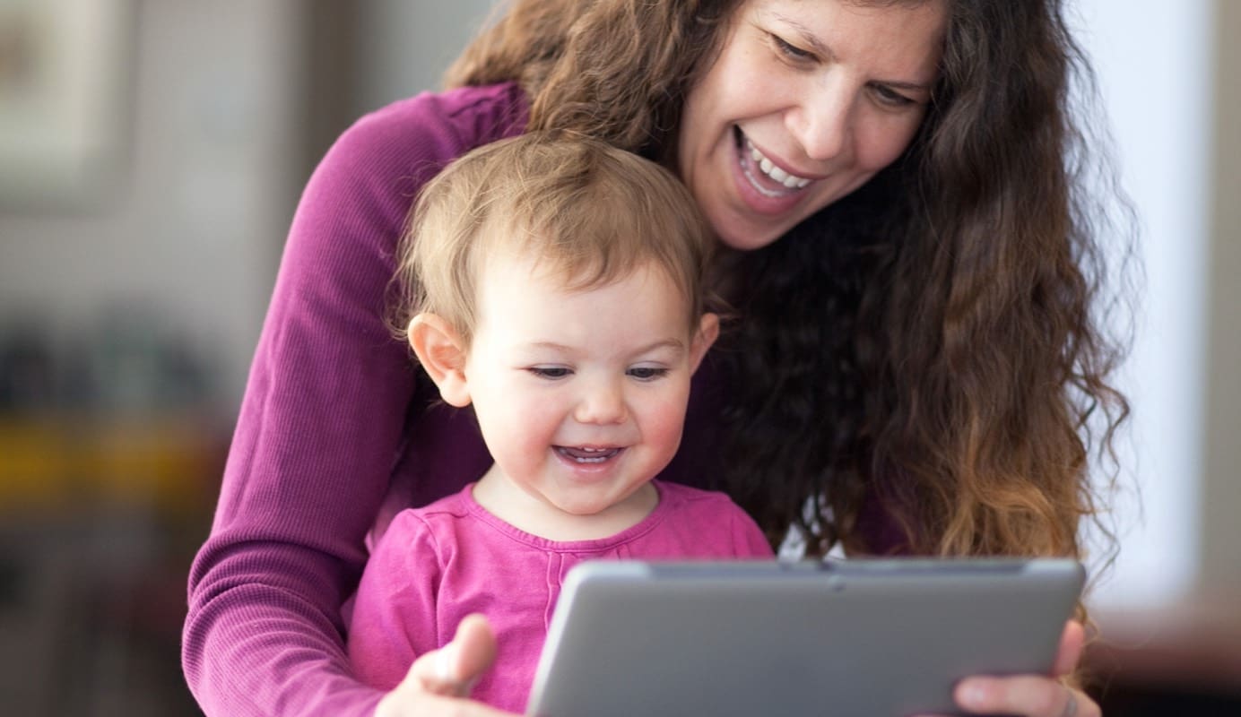 An adult and child smile cheerfully as they view a tablet that the adult is holding. Only the rear side of the tablet is visible.