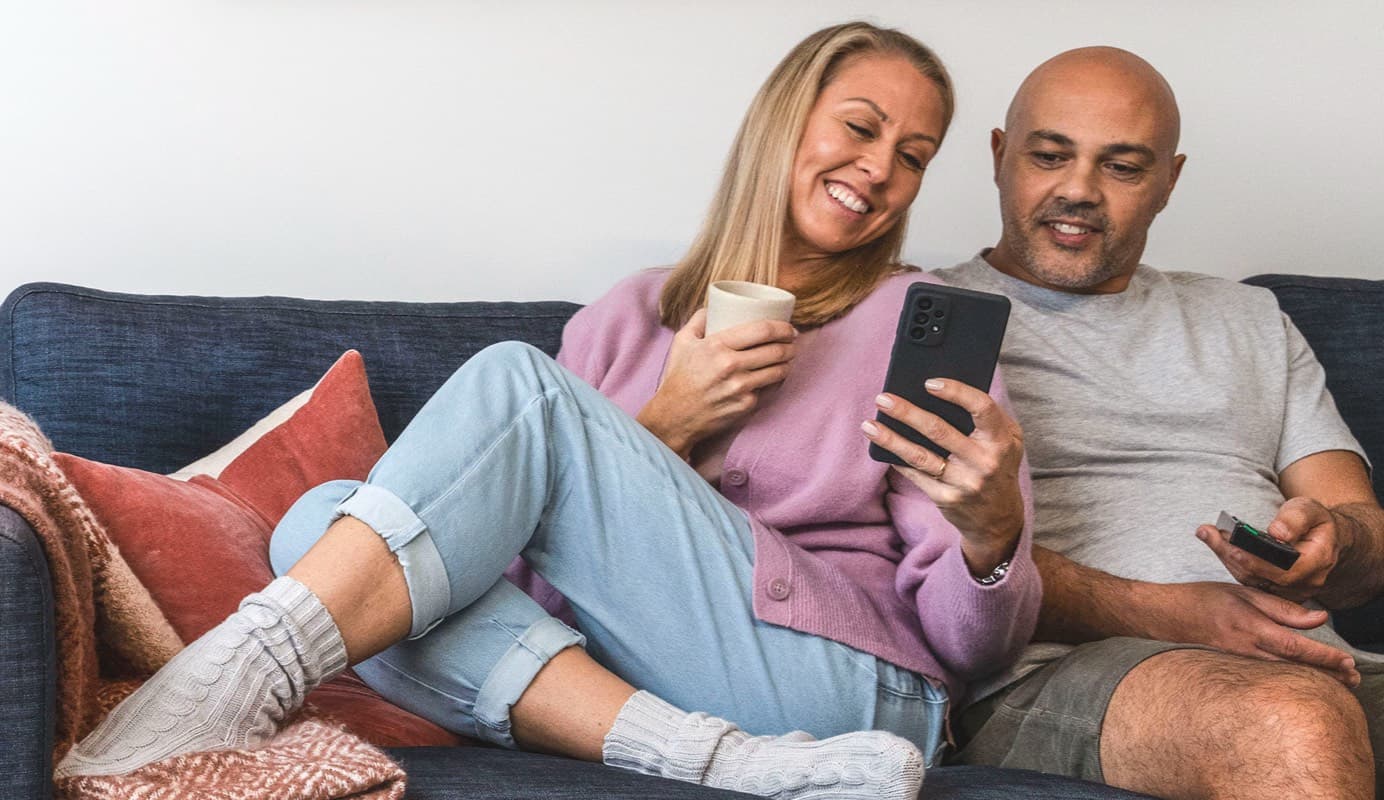 Two people smile at something on a phone screen while sitting together on a lounge.  