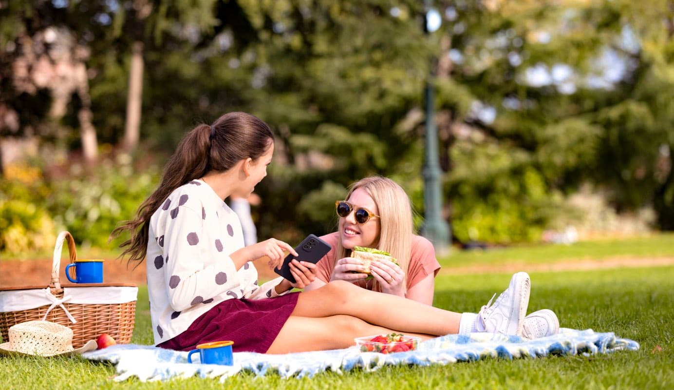 Two smiling adults enjoy a picnic and one is holding a mobile phone.