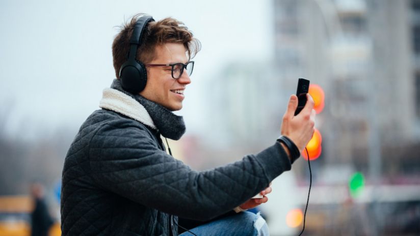 A man sitting outside in cold weather wearing a jacket and scarf smiles as he holds his smartphone up to make a video call.