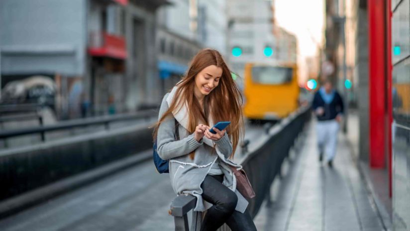 A woman waiting for public transport looks down at her smartphone and smiles.
